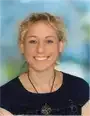 Eine Frau mit kurzen blonden Haaren lächelt strahlend. Sie trägt ein schwarzes Oberteil und eine Halskette mit Anhänger. Der Hintergrund ist unscharf und deutet darauf hin, dass sie sich in einem Raum befindet.
