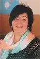 Eine lächelnde Frau mit kurzen schwarzen Haaren, die einen hellblauen Schal und ein schwarzes Oberteil trägt, posiert für die Kamera.