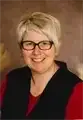 Eine Frau mit kurzen blonden Haaren, die schwarze Brille und ein rotes Shirt mit einem schwarzen Westchen trägt. Sie lächelt und scheint für ein Foto zu posieren.