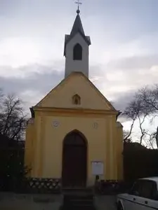 Eine kleine gelbe Kapelle mit einem Turm und einer Holztür, mit einem weißen Auto davor geparkt.