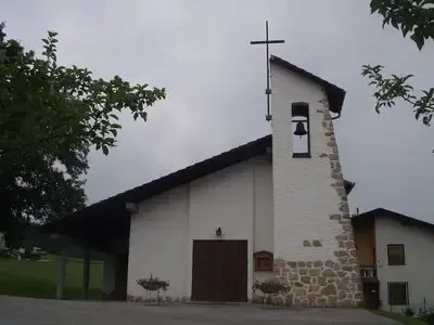 Eine weiße Kirche mit einem Kreuz oben, einem Glockenturm und einer braunen Tür ist von Bäumen und Gras umgeben.