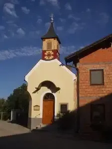 Eine kleine Kapelle mit einem spitzen Turm und einem Kreuz darauf, umgeben von Bäumen, unter einem blauen Himmel mit Wolken.
