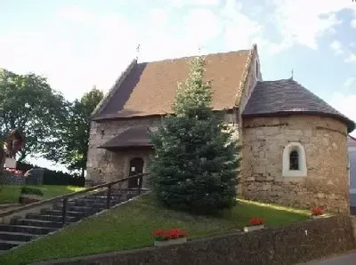 Eine alte Steinkirche mit einem braunen Dach und Stufen, die zu ihr hinaufführen. Ein Kiefernbaum und Blumen stehen vor der Kirche.