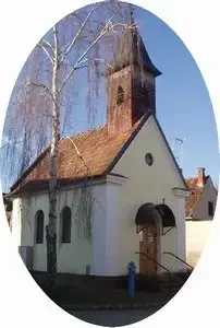 Eine kleine weiße Kirche mit einem braunen Dach und einem Turm steht vor einem Baum. Die Kirche hat eine braune Tür und ein Fenster.