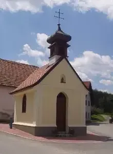 Eine kleine, weiße Kapelle mit einem roten Dach, einer Holztür und einem Kreuz oben, auf einer gepflasterten Fläche mit einem Gebäude dahinter.