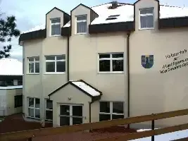 Ein weißes Gebäude mit mehreren Fenstern und einem kleinen Eingang. Schnee bedeckt das Dach und den Boden. Ein Holzzaun ist davor.