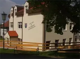 Ein beigefarbenes Gebäude mit einem roten Dach und Fenstern mit weißen Rahmen. Ein Holzzaun und Bänke stehen davor. Eine Straßenlaterne steht nebenan.
