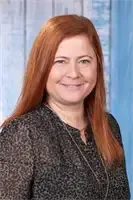 Eine Frau mit roten Haaren lächelt und trägt ein schwarzes Oberteil. Sie hat eine Halskette und steht vor einer blauen Wand.
