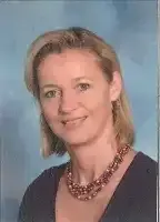 Eine Frau mit kurzen blonden Haaren lächelt und posiert für ein Foto. Sie trägt ein schwarzes Oberteil und eine Halskette mit braunen Perlen.