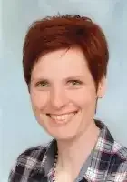 Eine Frau mit kurzen roten Haaren lächelt für ein Foto. Sie trägt ein kariertes Hemd in Blau und Weiß.