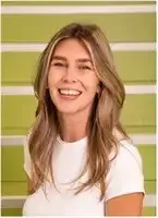 Eine Frau mit langen blonden Haaren lächelt fröhlich in die Kamera und steht vor einer grünen Wand. Sie trägt ein weißes T-Shirt und scheint für ein Foto zu posieren.