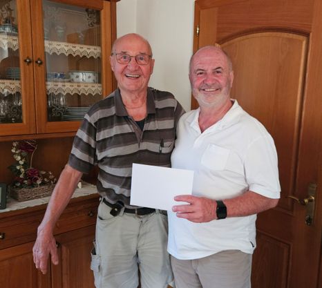 Zwei Männer stehen in einer Küche, einer hält ein Stück Papier in der Hand, beide lächeln. Hinter ihnen steht ein Holzschrank mit Glastüren, der Geschirr zeigt.