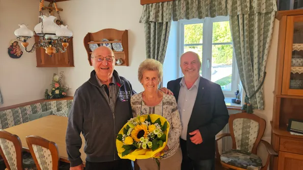 Drei Menschen stehen in einem Raum. Eine Frau hält einen Blumenstrauß.