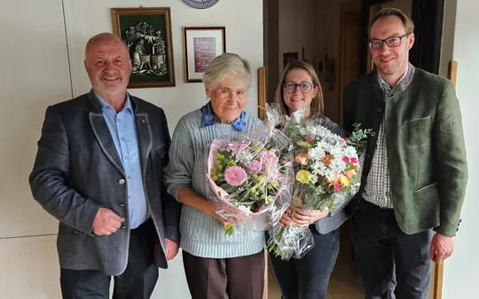 Eine Familie posiert für ein Foto vor einer weißen Wand mit Bilderrahmen, eine lächelnde ältere Frau hält Blumen.