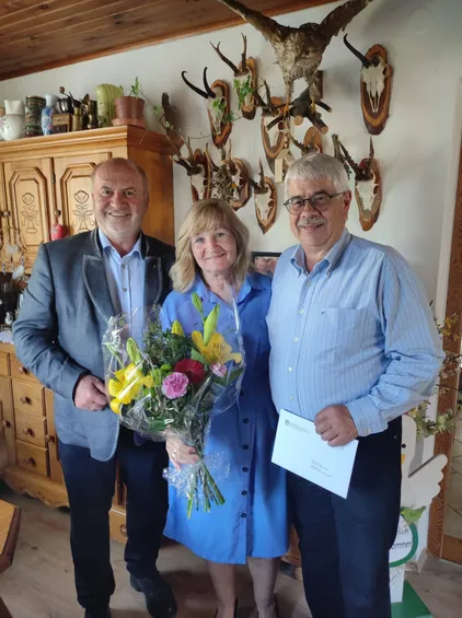 Drei Menschen stehen zusammen in einem Raum mit einem Blumenstrauß und lächeln für ein Foto. Dahinter befinden sich Trophäen und ein Holzschrank mit verschiedenen Gegenständen.