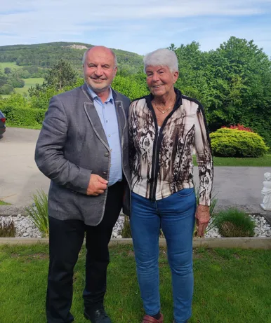 Ein älteres Paar steht auf einem Rasen mit malerischem Hintergrund. Der Mann trägt einen grauen Blazer und ein blaues Hemd, während die Frau eine gemusterte Bluse trägt. Sie lächeln beide in die Kamera.