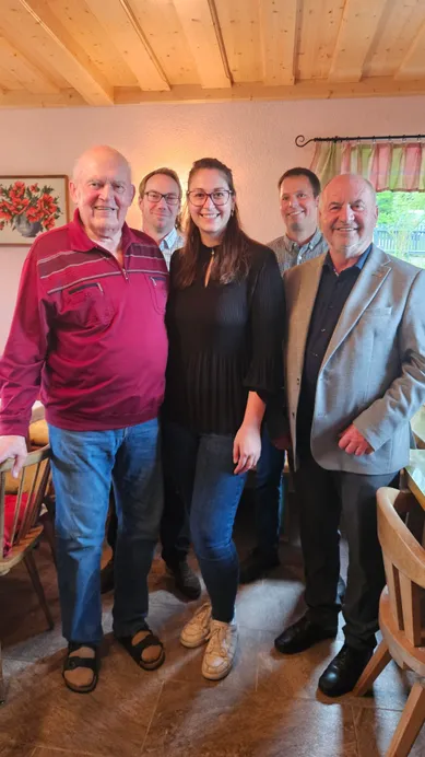 Fünf Menschen stehen lächelnd für ein Foto in einem Raum zusammen. Eine Frau in einer schwarzen Bluse steht zwischen zwei Männern. Hinter ihnen ist eine Wand mit einem Blumenbild.