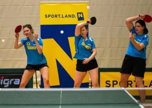 Drei Frauen spielen Tischtennis in einer Sporthalle. Die Spielerin in der Mitte schlägt den Ball mit ihrem Schläger. Dahinter ist ein Banner mit dem Wort Sport.land N und dem Logo.