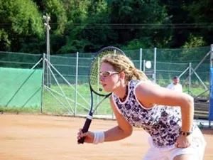 Eine Frau mit Brille und einer Uhr am linken Handgelenk spielt Tennis auf einem Sandplatz, während eine Person im Hintergrund zusieht.