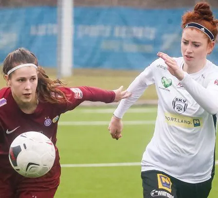 Zwei Fußballerinnen auf einem Spielfeld, eine in Rot mit einem Ball, die andere in Weiß schaut sie an.