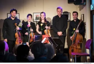 Fünf Musiker stehen vor einer weißen Wand, zwei mit Violinen, einer mit einem Cello und zwei mit Blumen. Eine der Geigerinnen lächelt, und es gibt ein Publikum vor ihnen.