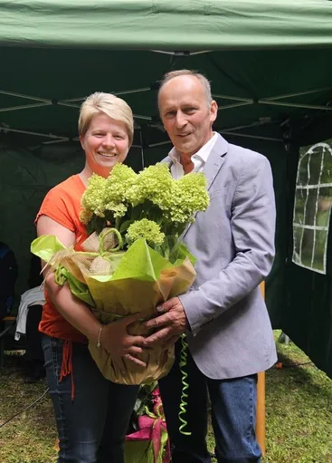 Ein Mann und eine Frau stehen unter einem Zelt und lächeln. Die Frau hält einen Strauß grüner Blumen. Der Mann trägt einen Anzug. Hinter ihnen steht ein Zelt mit einem Glasfenster.
