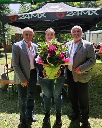 Drei Personen stehen unter einem Vordach mit einem Zeltschild, das '24MX' liest. Eine Frau hält einen Blumenstrauß. Hinter ihnen sind Tische und Stühle, mit sitzenden Menschen und anderen, die im Hintergrund gehen.
