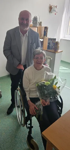Eine ältere Frau im Rollstuhl hält einen Blumenstrauß. Ein Mann in einem Anzug steht hinter ihr. Die Szene ist drinnen.