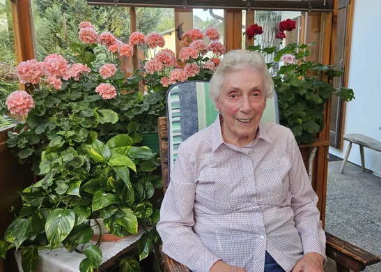 Eine ältere Frau sitzt auf einem Stuhl in einem Garten. Sie ist von leuchtend pinken und roten Blumen umgeben.