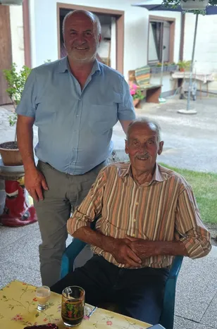 Zwei Männer sitzen draußen, einer in einem blauen Hemd, der andere in einem gestreiften Hemd, beide lächeln. Dahinter stehen ein Topf mit Pflanzen und eine Bank mit Blumen.