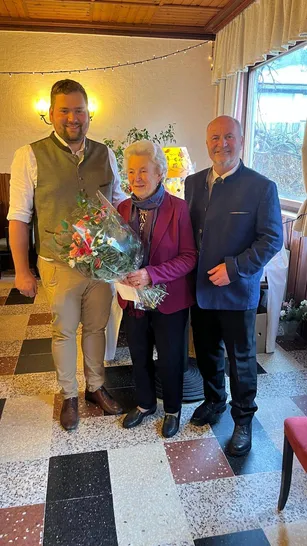 Drei Personen stehen in einem Raum. Eine Frau hält Blumen. Ein Mann links von ihr trägt eine Weste. Der Mann rechts von ihr trägt einen dunklen Anzug.