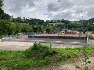 Ein leerer Tennisplatz, umgeben von einem hohen Zaun und Bäumen, mit einem bewölkten Himmel darüber.