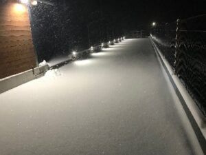 Eine schneebedeckte Straße mit einem Holzgebäude links, einem Zaun rechts und beleuchteten Straßenlaternen.