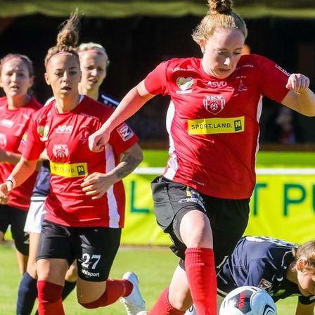 Ein Fußballspiel mit Spielerinnen in roten und blauen Trikots auf einem Spielfeld, eine Spielerin in Rot jagt den Ball, während eine andere in Blau am Boden liegt. Ein Banner dahinter lautet 'L Pure'.