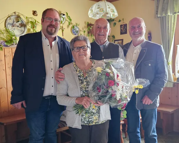 Vier Personen stehen in einem Raum und lächeln. Eine Frau hält einen Blumenstrauß und drei Männer stehen hinter ihr, alle in Anzügen gekleidet. Die Wand ist mit Tellern und Pflanzen dekoriert.