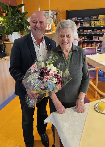 Ein Mann und eine Frau stehen in einem Raum und lächeln. Der Mann hält einen Blumenstrauß. Sie stehen in der Nähe eines Tisches mit einem Glas Orangensaft. Dahinter befindet sich ein Regal mit Snacks.