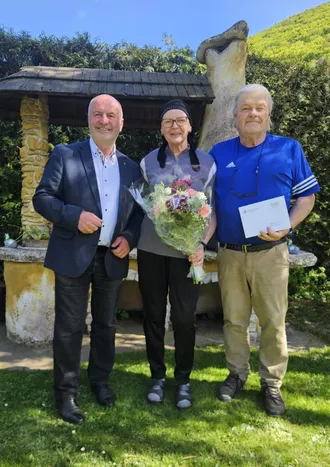 Drei Menschen stehen in einem Garten zusammen. Eine Frau hält einen Blumenstrauß. Ein Mann auf der rechten Seite hält ein Zertifikat. Dahinter steht ein Steinpavillon.