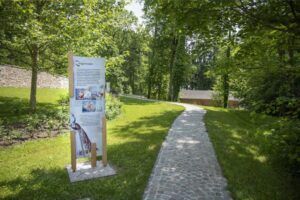 Ein Schild steht in einem Rasenbereich mit einem Steinweg, der zu einem Gebäude führt. Bäume umgeben den Bereich. Das Schild hat Bilder von Menschen und Tieren.