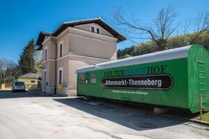 Ein grüner Zugwaggon mit der Aufschrift 'Kulturrainhof Altenmarkt-Zhennerberg' steht am Straßenrand mit einem Gebäude im Hintergrund.