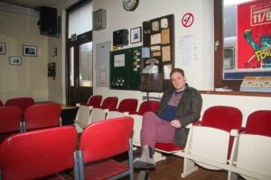 Ein Mann sitzt auf einem roten Stuhl in einer Wartehalle. Er trägt eine Jacke und hält eine Tasche. Der Raum hat weiße Wände und rote Stühle.