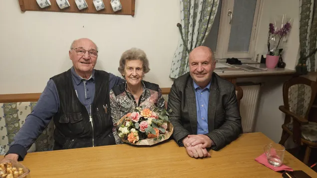 Drei Personen sitzen um einen Tisch mit einem Blumenstrauß. Der Mann auf der linken Seite trägt eine Brille und eine Weste. Die Frau in der Mitte lächelt und trägt eine Bluse mit Blumenmuster. Der Mann auf der rechten Seite trägt einen Anzug und hat seine Hände ineinander verschränkt. Hinter ihnen befindet sich eine Wand mit einem Fenster und Vorhängen.