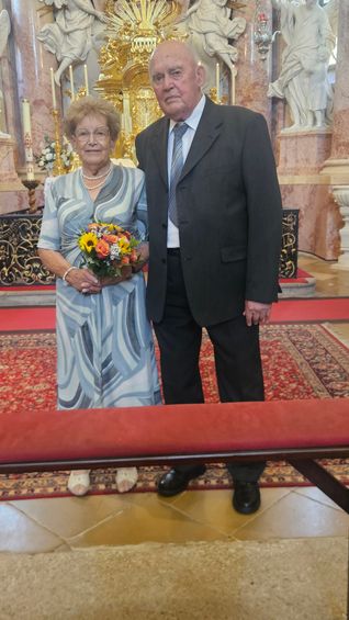 Ein älteres Paar steht in einer Kirche, die Frau hält einen Blumenstrauß, beide lächeln und schauen zur Kamera.
