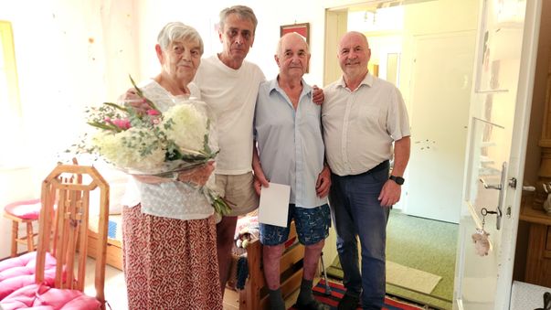 Drei ältere Personen stehen zusammen in einem Raum, eine Frau hält einen Blumenstrauß. Der Mann in der Mitte hält ein Stück Papier.