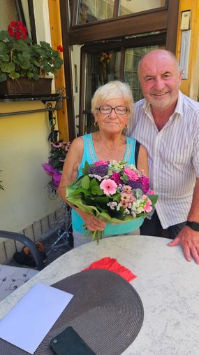Eine ältere Frau mit Brille und einem blauen ärmellosen Oberteil hält einen Blumenstrauß in der Hand, während sie neben einem lächelnden Mann in einem gestreiften Hemd steht.