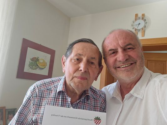Zwei Männer posieren für ein Foto, einer hält ein Zertifikat und der andere lächelt. Hinter ihnen ist eine Wand mit einem gerahmten Bild und einer Uhr.