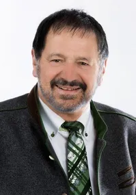 Ein Mann in Anzug und Krawatte, der eine Weste mit grün-braunen Besätzen trägt, lächelt in die Kamera.