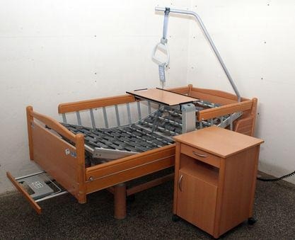 Ein Krankenhausbett mit Metallrahmen und hölzerne Kopfstütze steht an einer weißen Wand. Es verfügt über eine ausziehbare Ablage und einen Nachttisch. Der Raum ist leer, mit einem grauen Teppichboden.