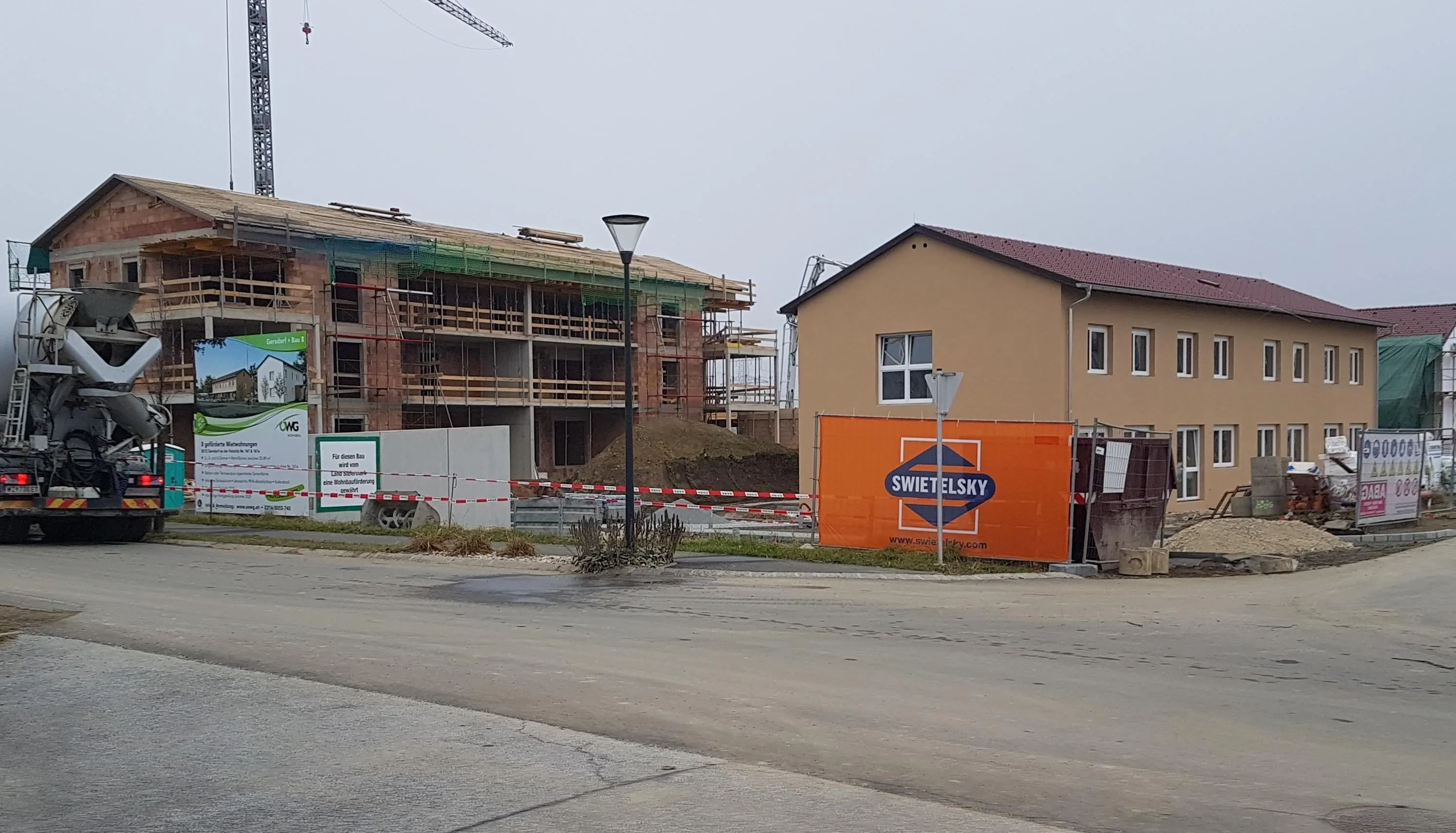 Eine Baustelle ist im Gange mit einem großen Gebäude im Bau. Ein fertiggestelltes Haus mit rotem Dach steht rechts. Ein Straßenlicht steht am Straßenrand.