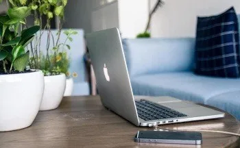 Ein Apple-Laptop steht auf einem Holztisch mit Topfpflanzen und einem blauen Sofa im Hintergrund. Ein Telefon ist an den Laptop angeschlossen.