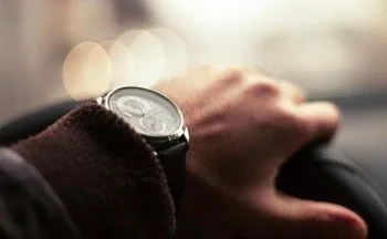 Eine Person in einer braunen Jacke hält ein Lenkrad mit einer Uhr am Handgelenk, mit verschwommenen Stadtlichtern im Hintergrund.